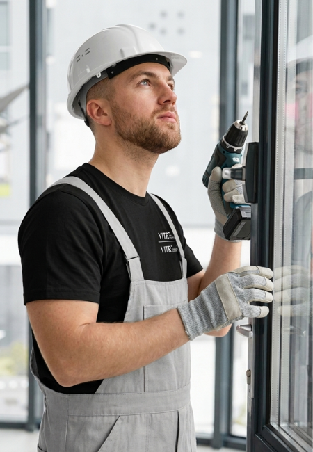 Técnico instalador con casco y taladro ajustando una puerta de aluminio y vidrio de alta calidad.